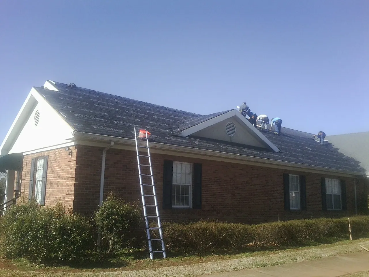 Crew tearing shingles off a brick ranch roof with a ladder set — Emergency Roof Repair work by our crew in Hazlehurst
